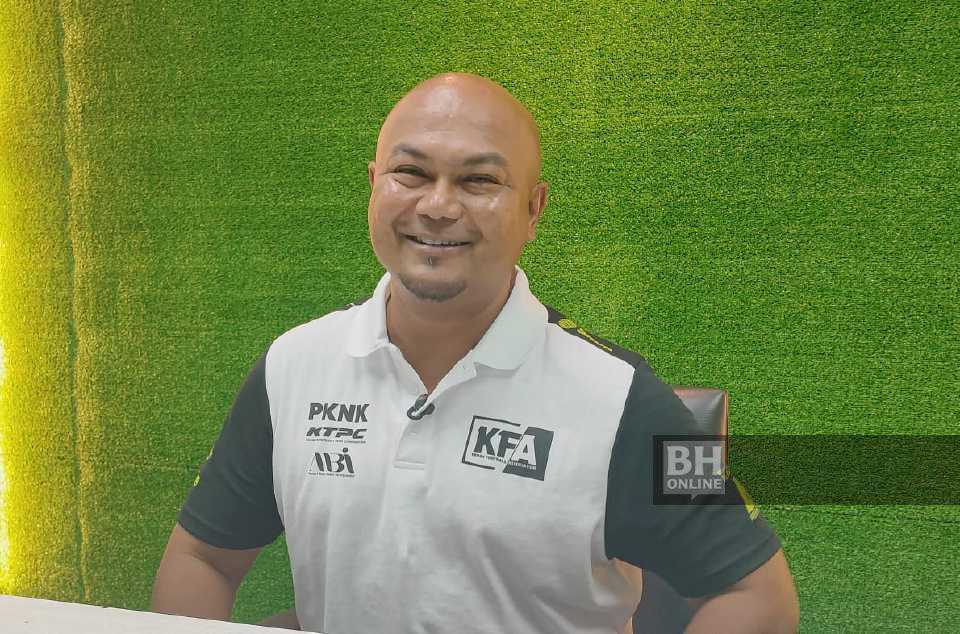 Akmal Rizal smiling at a press conference in a white Kedah FA polo shirt with dark sleeves, the club crest on his chest beside the PKNK, KTPC and ABI sponsor marks, against a backdrop of green artificial turf